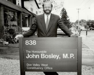 Image shows a politician John Bosley standing behind a sign marking his riding office. 