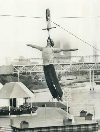 Hanging by the back of his neck, stuntman Jay Cochrane executes a daring slide from the pod roof of Ontario Place to the reflecting pool on the west i(...)