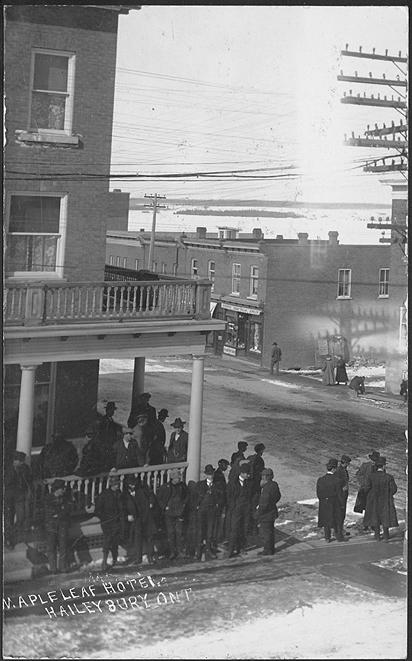 A large crowd of men stand outside a three story hotel building with a balcony.