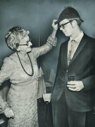 Shows True Davidson tweaking the brim of a Sherlockian Deerstalker hat worn by a tall young man…