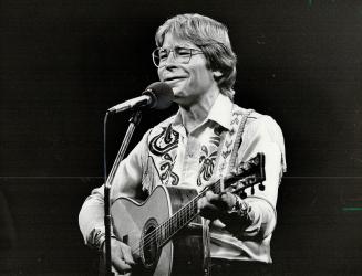 A smiling blonde man, wearing an embroidered shirt and large glasses, plays an acoustic guitar  ...