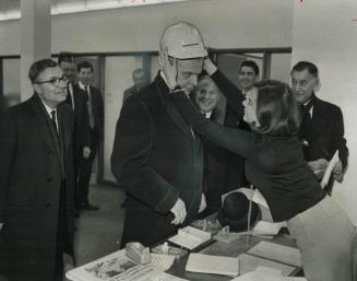 Dief Dons hard hat: Secretary Mrs. Marilyn Devanney helps Opposition Leader John Diefenbaker on with a safety helmet before he goes out to tour Expo '(...)