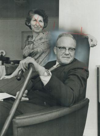 A man, holding a cane, sits in an armchair on which perches a dark-haired woman. Both are smili…