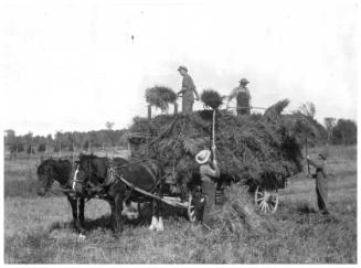 Farm workers with pitchforks loading hay onto a horse-drawn wagon.
