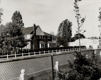 Side view of brick home seen over the top of a chain link fence, in foreground. Trees are seen  ...