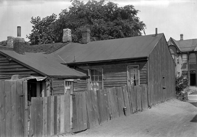Shows a rickety back fence running along two wooden buildings in poor repair, with tar-paper ro…