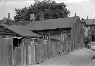 Shows a rickety back fence running along two wooden buildings in poor repair, with tar-paper ro…