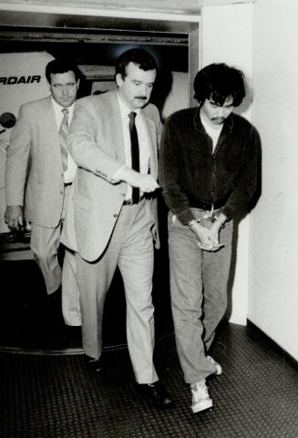 A young Asian man in handcuffs, is escorted by two large White men in suits and ties. In the ba ...