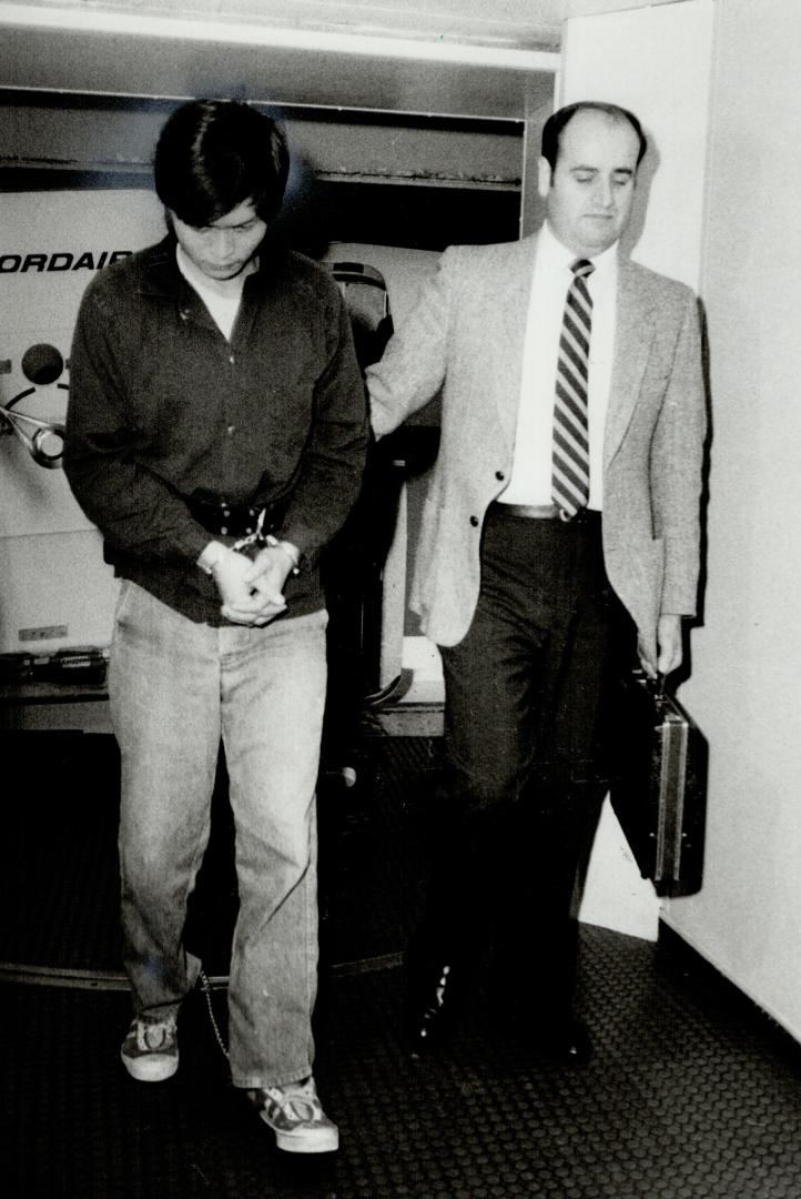 A young Asian man in handcuffs and wearing a chain on his ankles is escorted by a taller man in ...