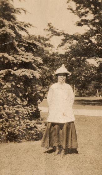 In the foreground, a woman in a light-coloured hat and shirt and long, dark dress stands smilin ...