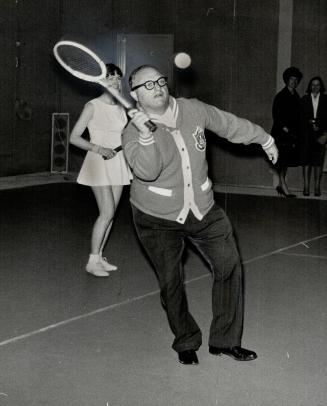 In which he serves. Mayor Givens is backed up by Mrs. H. Goulding as he opened York Racquets Club on Yonge St., Toronto's first club built specifically for indoor tennis