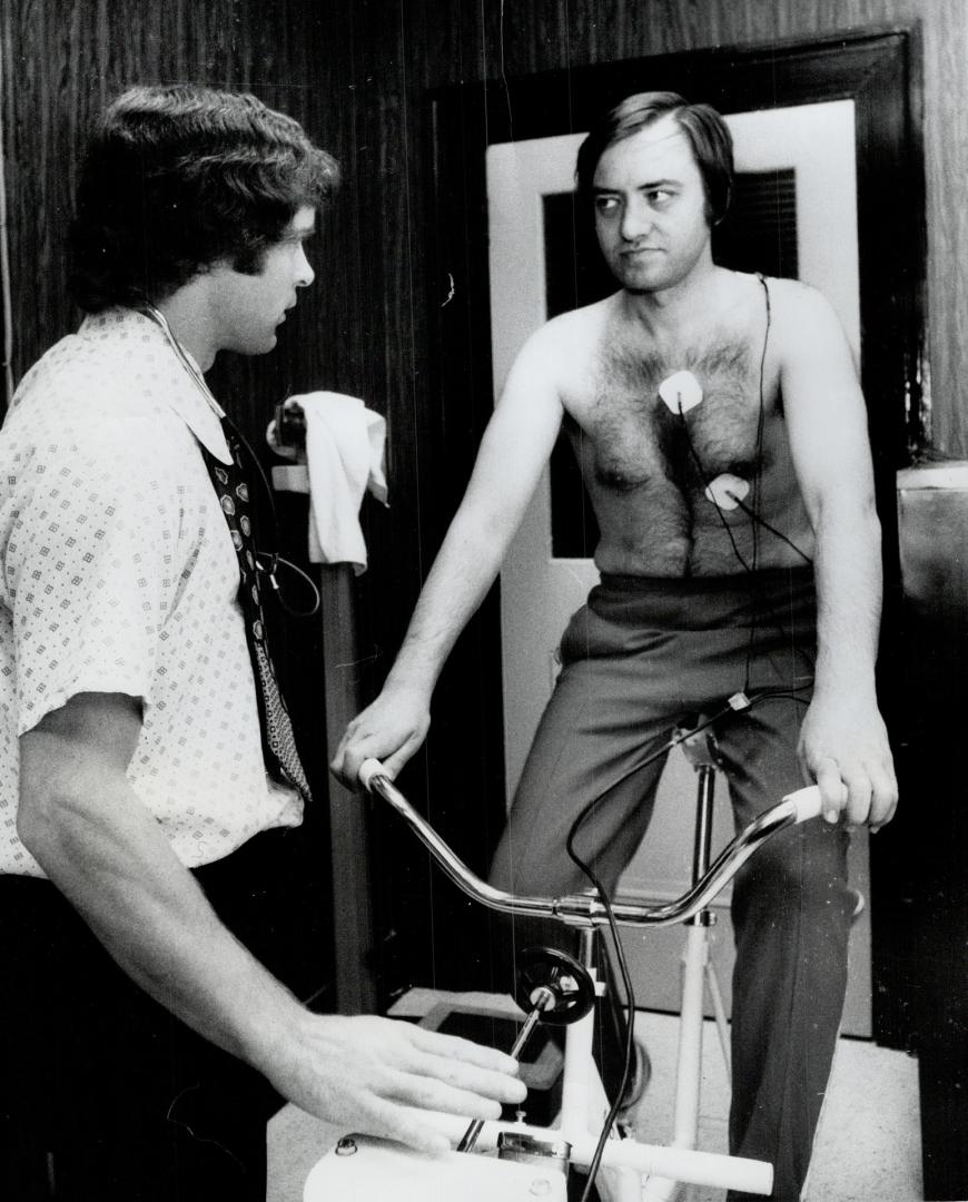 Metro Chairman Paul Godfrey takes his sweat suit off and mounts cycle machine as he is tested for general physical fitness by Don Payne, Associate Director of Physical Fitness at Central Y