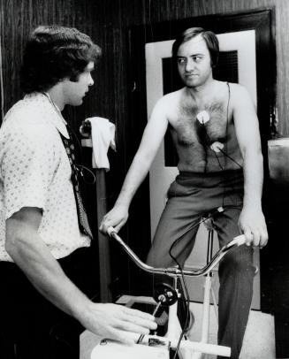 Metro Chairman Paul Godfrey takes his sweat suit off and mounts cycle machine as he is tested for general physical fitness by Don Payne, Associate Director of Physical Fitness at Central Y
