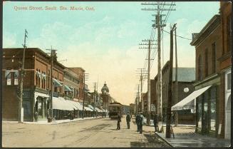 A lively street scene. A group of men stand on the unpaved roadway, a streetcar heads away from…