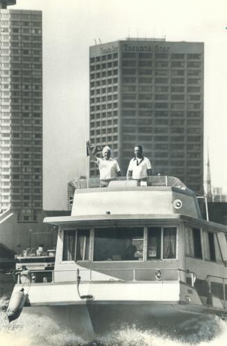 Shows a houseboat leaving Toronto harbour with the Toronto Star building in the background. Two ...