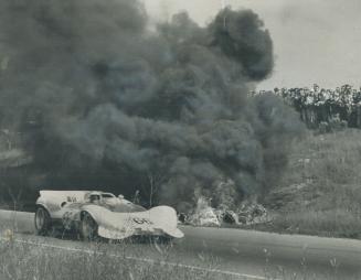 Grand prix winner Jim Hall whizzes past pall of black smoke from burning car: Crowd gathers above track to watch Herb Swann's Elva-BMW reduced to ashes on 11th lap of 250-mile classic at Mosport