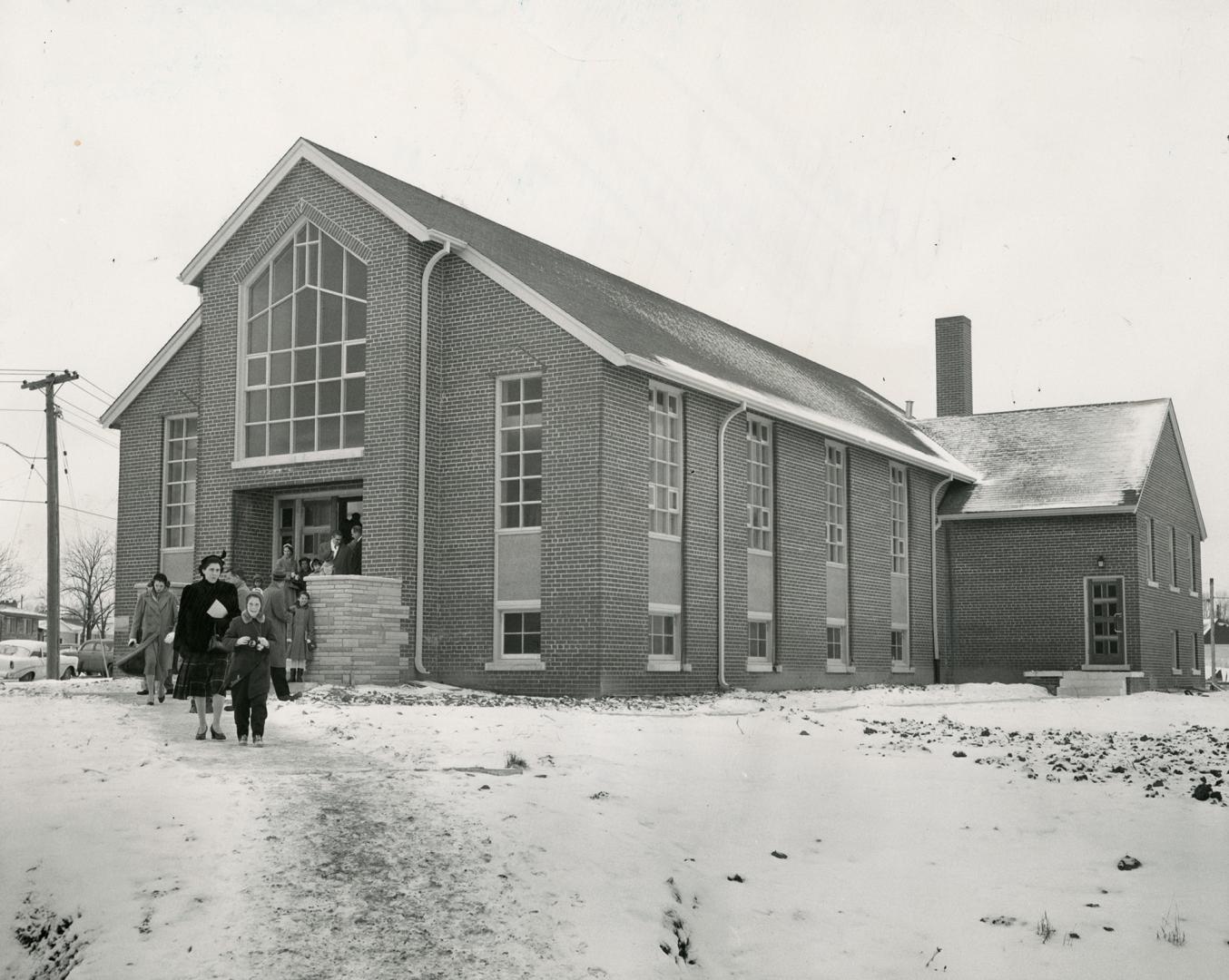 Small crowd seen exiting gable-roofed church with lattice window above entrance.