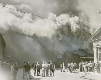 People stand in the street watching thick, billowing smoke that fills the sky. A firehose is vi ...