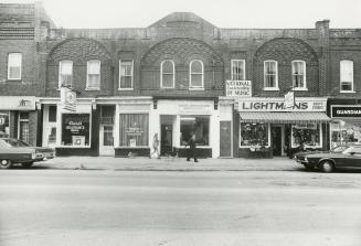 Shows ornate brick storefronts. A couple and a dog walk along the sidewalk. 1970s-era cars are  ...