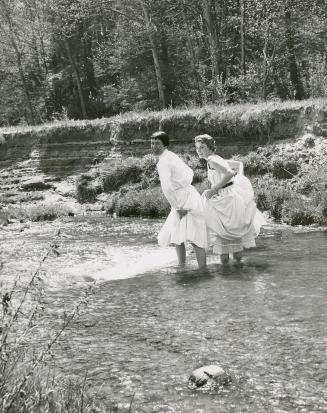 Two women in long white dresses wade in shallow waters of river; treed shore in background.