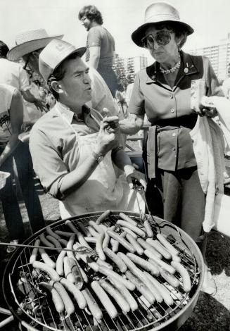 A man in a Liberal baseball cap stands in front of a barbecue grill full of hotdogs, while he s ...
