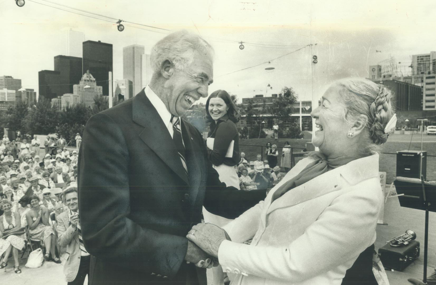Image shows a gentleman and a lady shaking hands and laughing on stage with the audience in the…