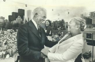 Image shows a gentleman and a lady shaking hands and laughing on stage with the audience in the…