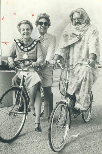 There's a reason. Alderman Frank Bean is dressed in wig and dress. It's to promote women's bike-a-thon May 6 to aid cancer society. Mayor Hazel McCallion rides cross bar on MPP Bud Gregory's bike