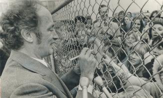 Giving an autograph through a wire fence is quite a trick, but Prime Minister Pierre Trudeau does it with pleasure at the Saskatoon airport