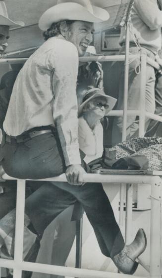 Perched casually on a fence, Prime Minister Pierre Trudeau watches roping, riding and steer-wrestling at the Calgary Stampede and riding at the Calgary Stampede where 25,000 spectators cheered him