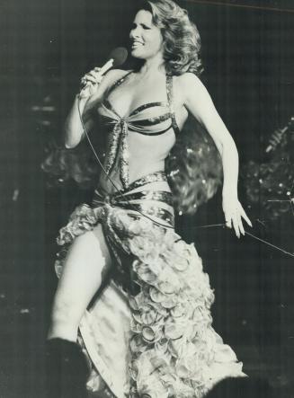 Shows a woman in a dress with a bikini top and a high-cut skirt singing into a microphone.