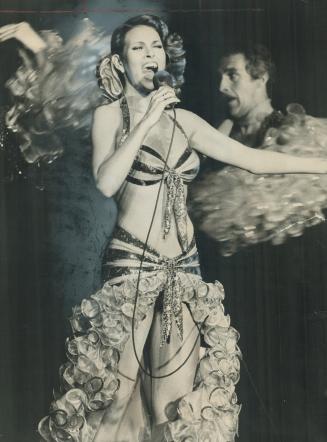 Shows a woman in a bikini top and high-cut skirt singing enthusiastically into a microphone, wh…