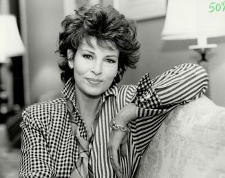 A woman with short curly hair smiles at the camera while sitting on a chesterfield in a living …