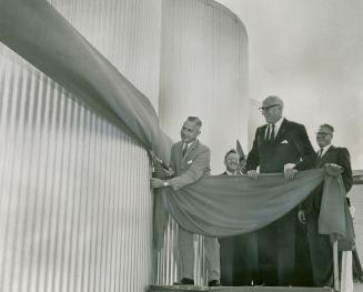 Man in suit cuts ribbon surrounding large, corrugated, columnar structure using oversized sheer ...