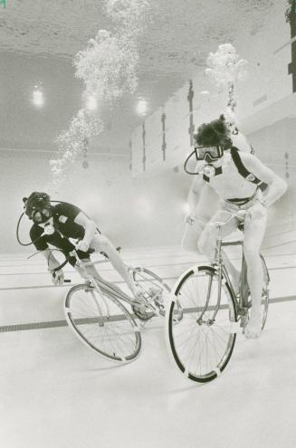 Two barefoot men ride bikes underwater in a white tiled pool; they wear scuba tanks strapped to ...