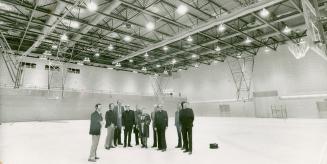 Small group of men and one woman stand in brightly lit empty gymnasium, looking in direction of ...