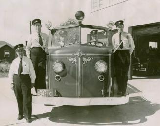 Four firemen in white shirts, black pants, ties and peaked officer caps, pose with fire engine. ...