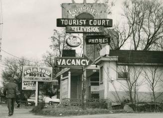Collection of signs staked into lawn and attached to pole beside a low, white building with bla…