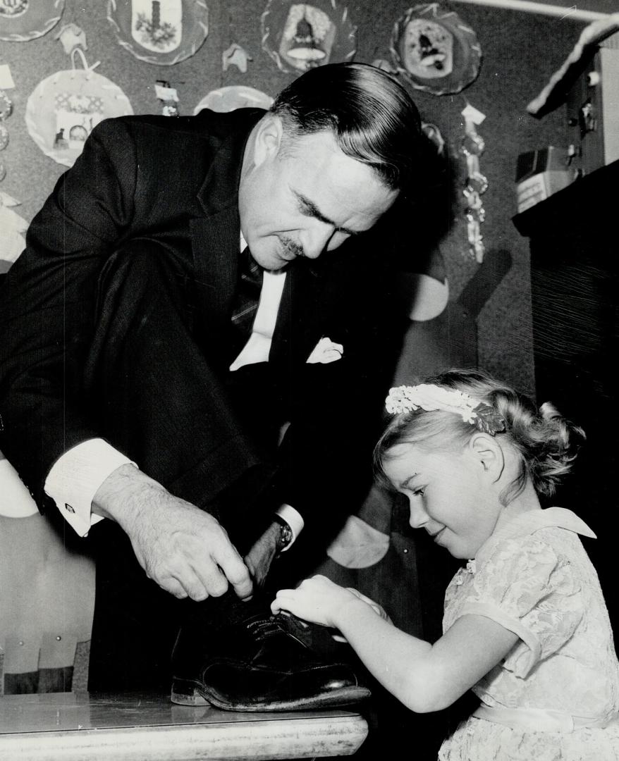 Helping hand. Mary Miles, a pupil at the Beverley St. School for Retarded Children, demonstrates her skill at tying shoe laces on Premier John Robarts(...)