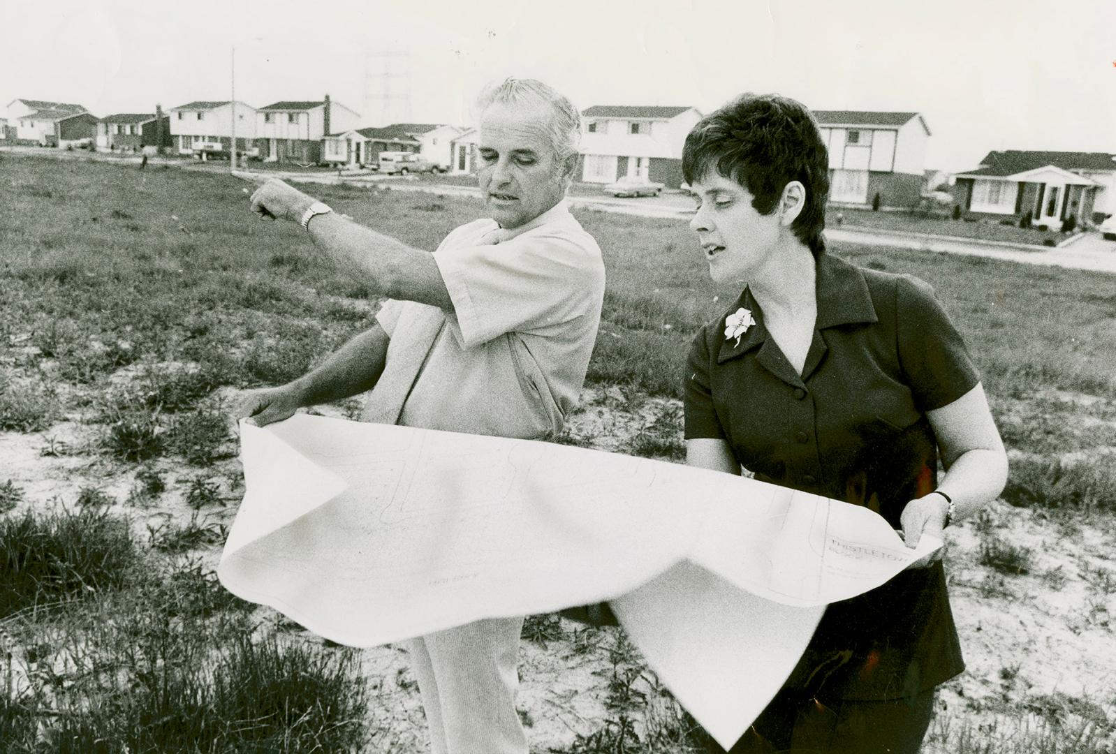 Man and woman stand on open grassy land examining large sheet of paper held between them; man p…