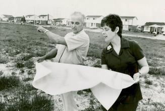 Man and woman stand on open grassy land examining large sheet of paper held between them; man p…