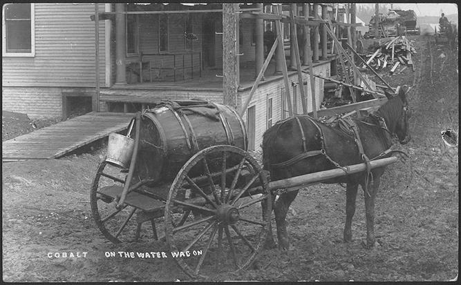 Black and white photograph of a horse pulling a wagon with barrel in front of a building with a ...