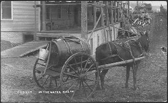 Black and white photograph of a horse pulling a wagon with barrel in front of a building with a ...