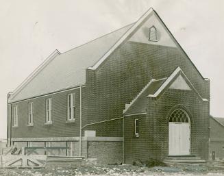 Brick church with stone foundation, steeply pitched roof and covered entrance. Wood planks pile ...
