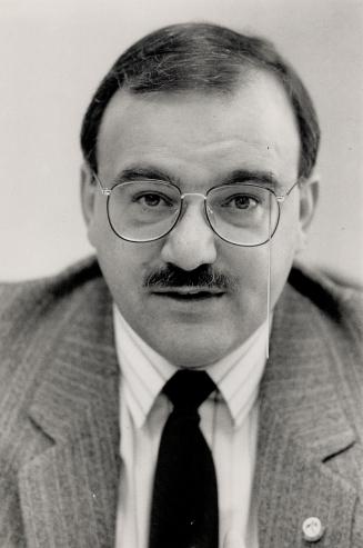 A close-up of a mustached man, with a pin-striped suit and round glasses, looking into the came ...