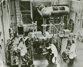 Three men in white lab coats look at various pieces of very large equipment.