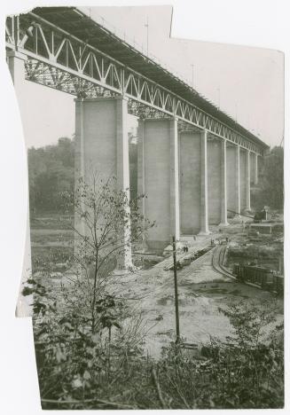 Underside of viaduct showing concrete piers supporting steel rails. At ground level below, smal ...