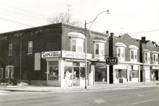 A corner shot of three duplex-style storefronts, the left-most of which is Tom's Variety Grocer ...