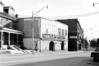 A two-storey square white brick building with very few windows. Two large signs over two arched…