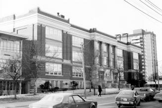 Shows a large school building with three storeys. It has columns and trees, iron fencing. There…